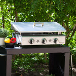 Plancha inox à trois brûleurs avec couvercle relevé en extérieur, posée sur un chariot noir, entourée de verdure et de légumes frais.