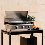 Plancha inox à trois brûleurs avec couvercle relevé, posée sur un chariot noir devant un mur clair, accompagnée de légumes et d’une plante décorative.