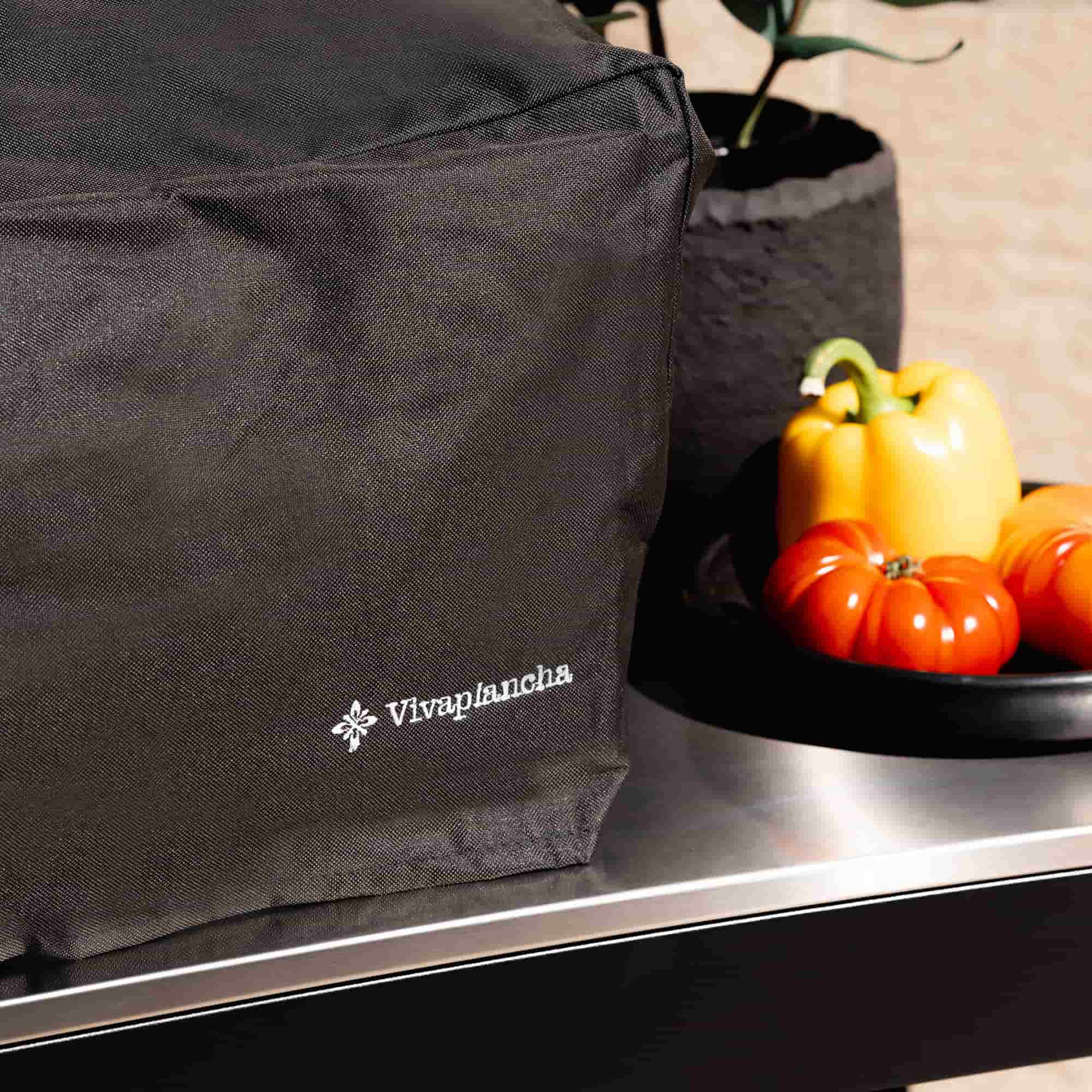 Gros plan sur la housse noire Vivaplancha couvrant une plancha, avec logo visible au premier plan, posée sur une desserte inox et entourée de légumes.