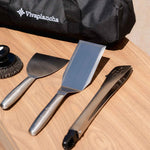 Ustensiles Vivaplancha posés sur un plan de travail en bois : spatule large, spatule longue, pince en inox et boule de nettoyage, avec le sac de transport visible en arrière-plan.