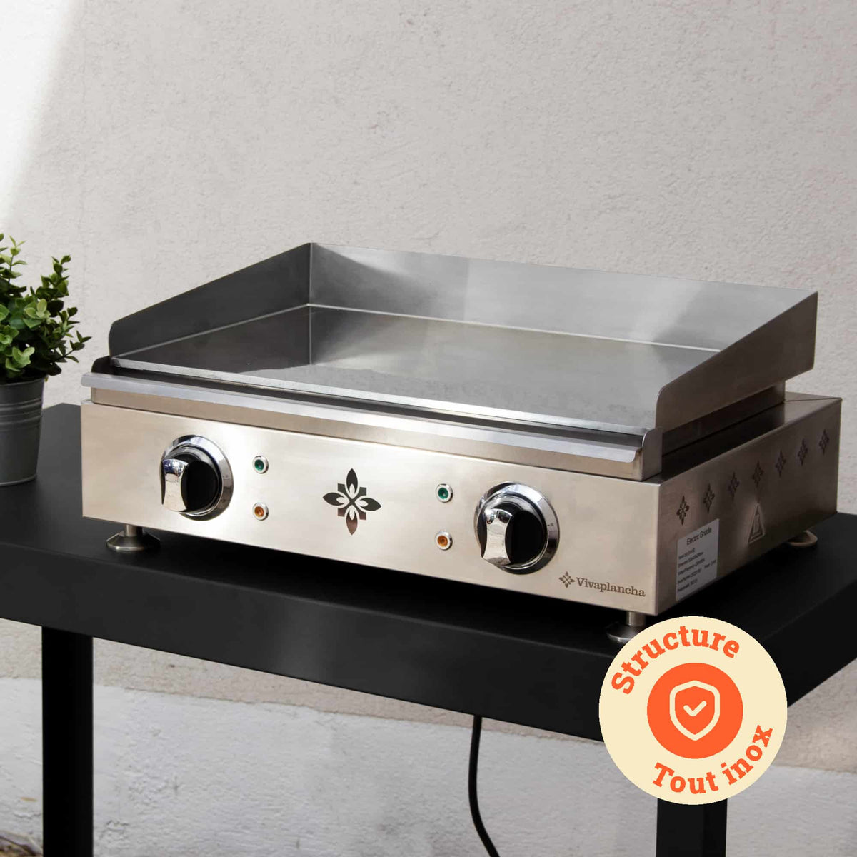 Vivaplancha plancha geplaatst op een buitentafel, met de volledig roestvrijstalen structuur in de kijker en een badge “Volledig roestvrijstalen structuur” weergegeven in een oranje cirkel.